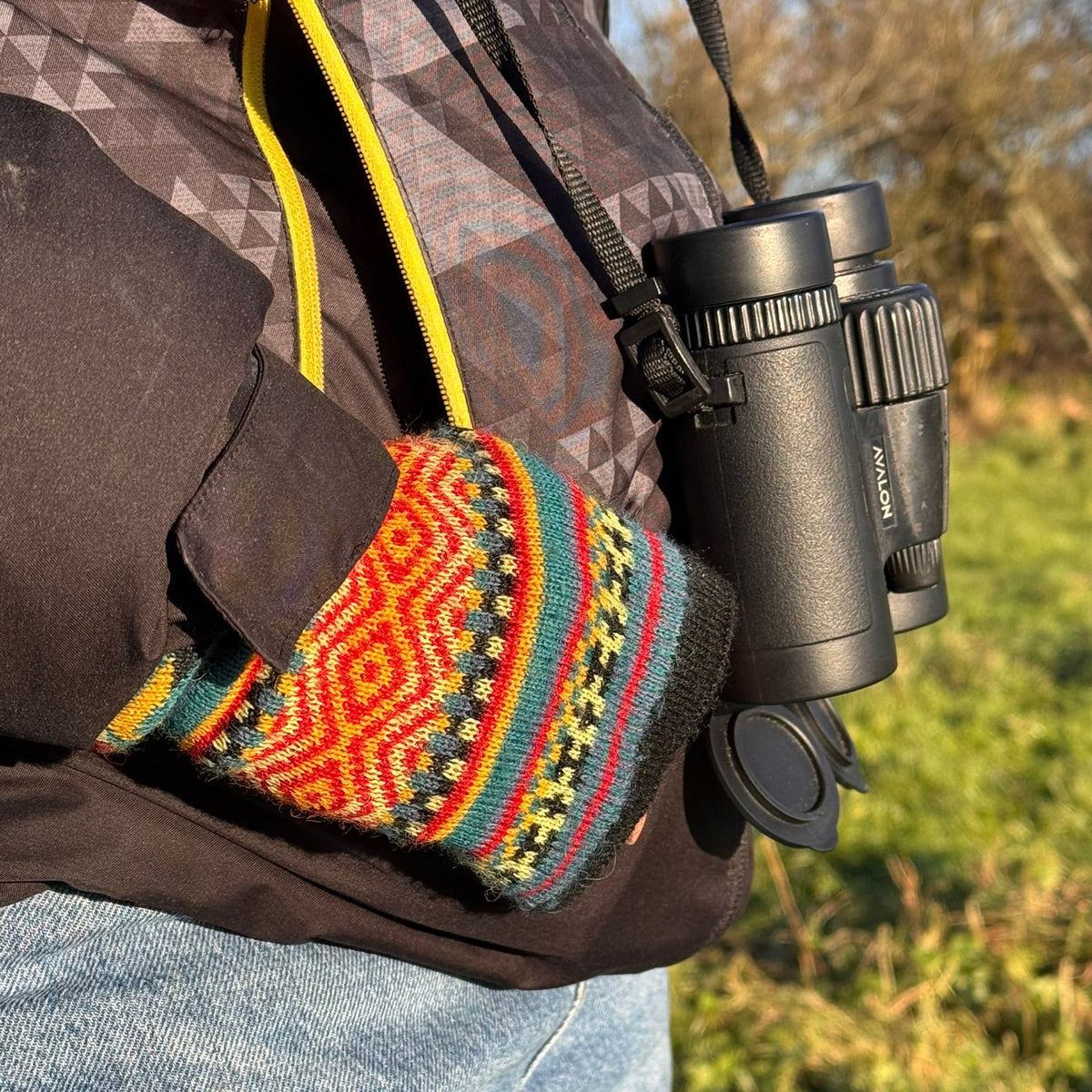 A close-up lifestyle shot of a person outdoors wearing a brown jacket and blue jeans, holding a pair of black binoculars. A colorful knit cuff with a red, orange, yellow, green, and blue geometric pattern is wrapped around the person's wrist.
