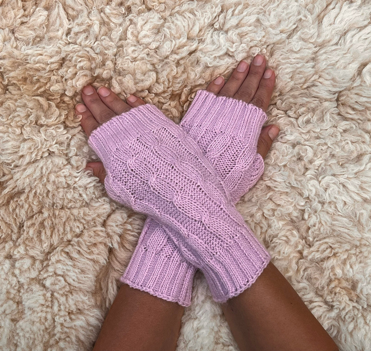 Pale Lilac Pink Cable Knit Alpaca Fingerless Gloves worn on hands resting on a sheepskin rug