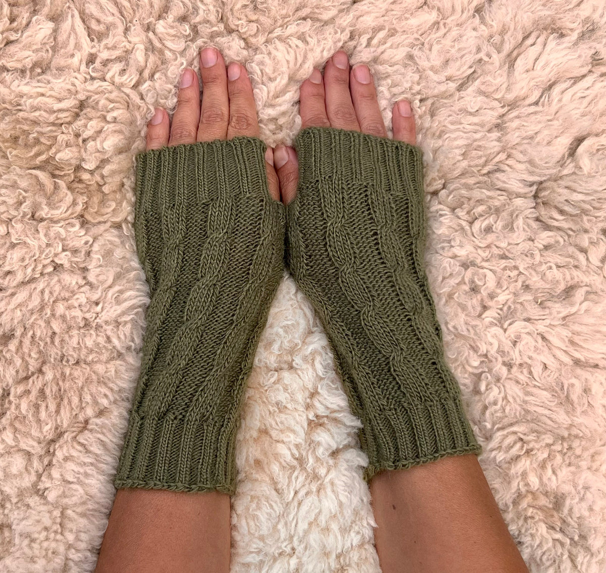Olive Green Cable Knit Alpaca Fingerless Gloves worn on hands resting on a sheepskin rug