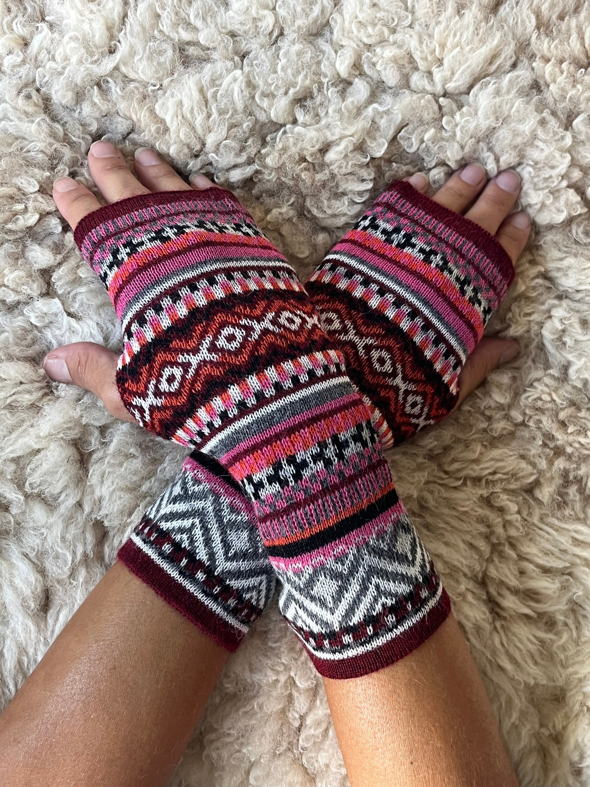 Burgundy & Pink Fair Isle Baby Alpaca Fingerless Gloves worn on hands resting on a sheepskin rug