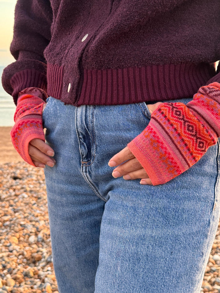 Coral and burgundy Fair Isle gloves, hands in pockets on the beach