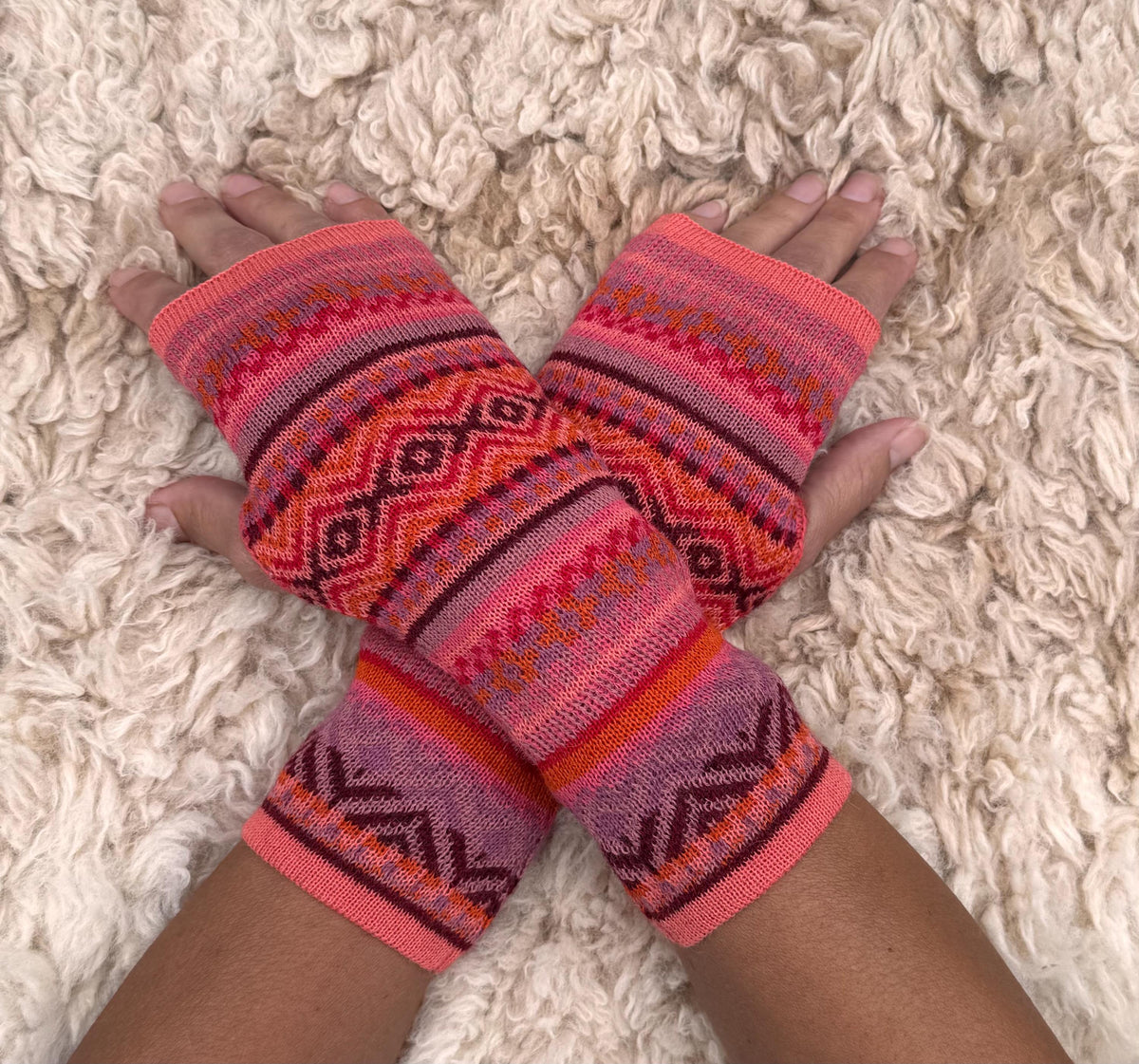 Coral & Burgundy Fair Isle Baby Alpaca Fingerless Gloves worn on hands resting on a sheepskin rug