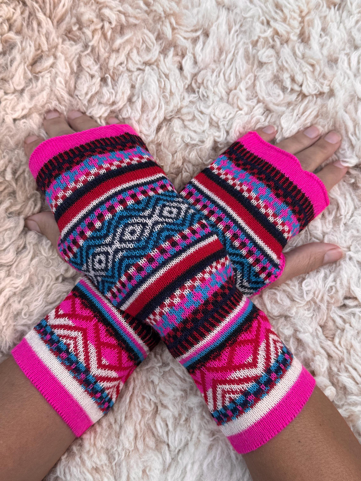 Neon Pink & Teal Fair Isle Baby Alpaca Fingerless Gloves worn on hands resting on a sheepskin rug