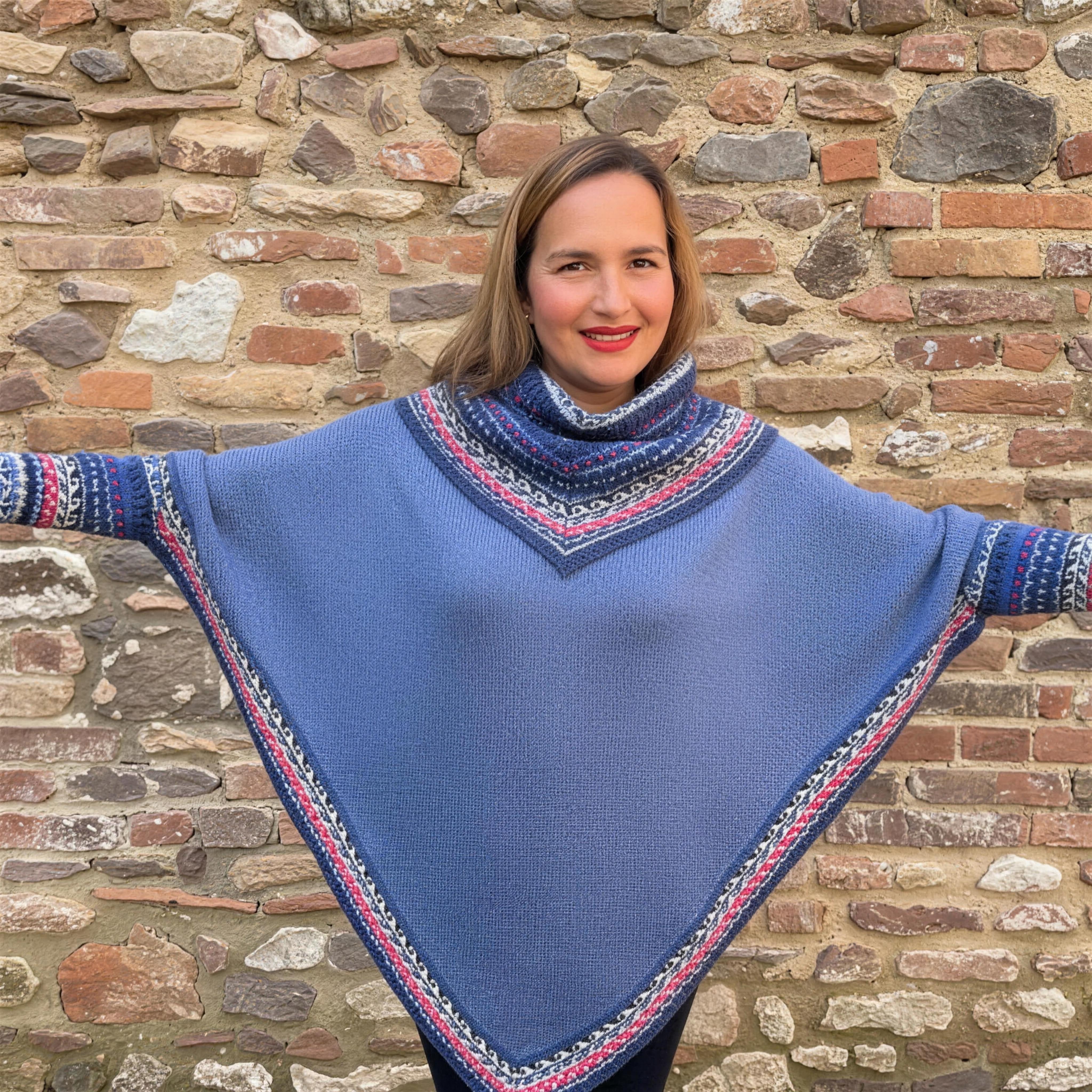 A woman with her arms out in a blue alpaca poncho with Fair Isle borders, against a flint and brick wall