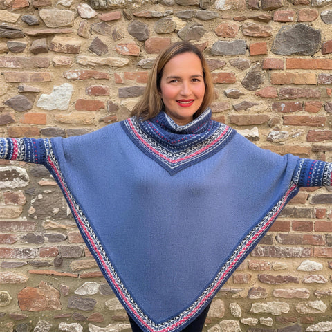 A woman with her arms out in a blue alpaca poncho with Fair Isle borders, against a flint and brick wall