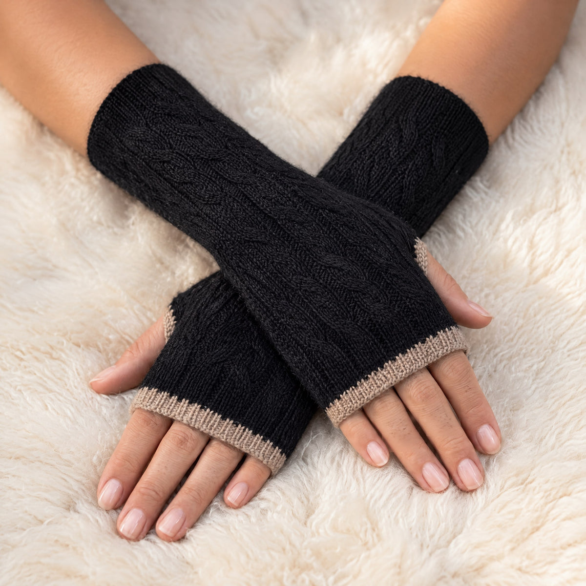 Black & Camel Reversible Cable Knit Baby Alpaca Fingerless Gloves worn on hands resting on a sheepskin rug