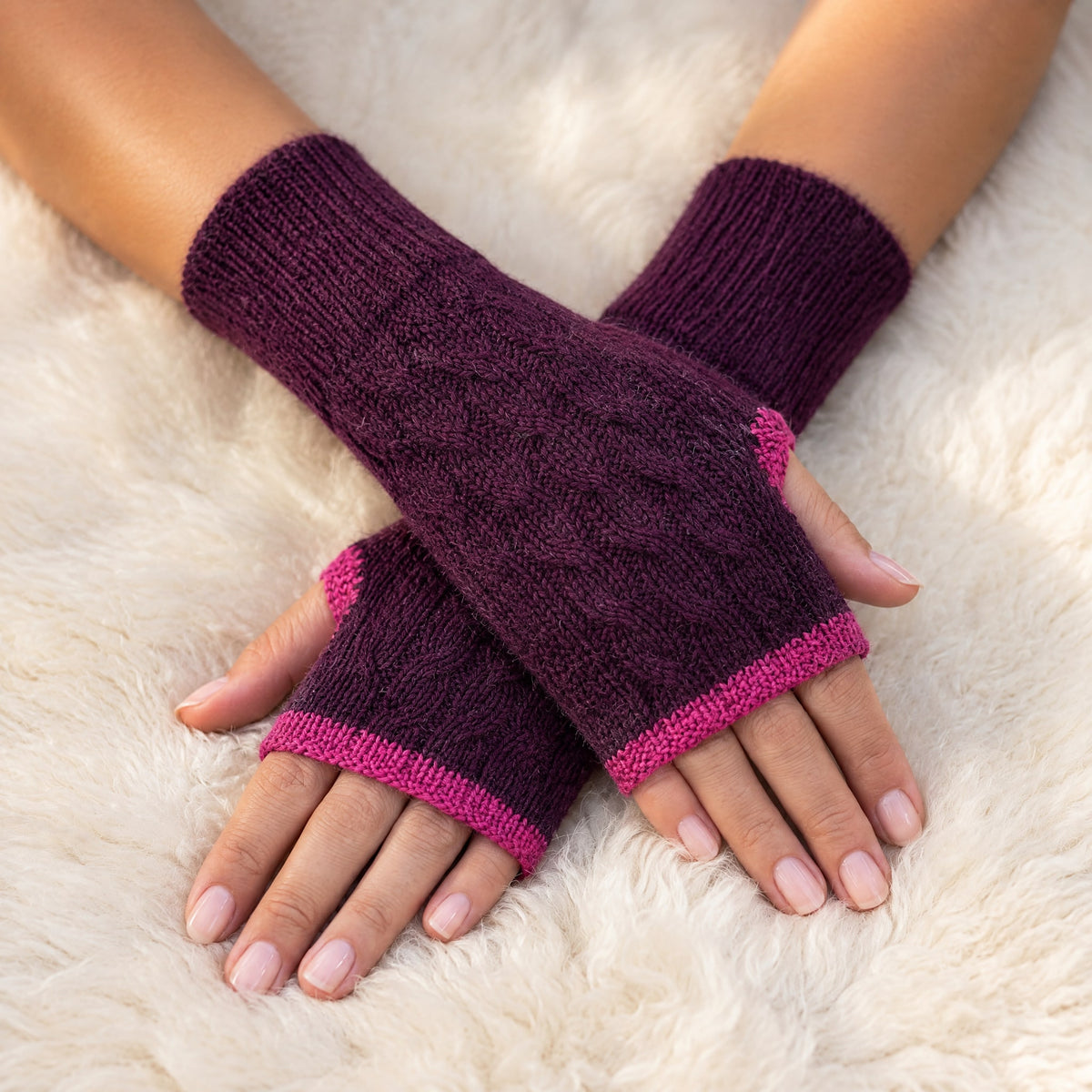 Burgundy & Pink Reversible Cable Knit Baby Alpaca Fingerless Gloves worn on hands resting on a sheepskin rug