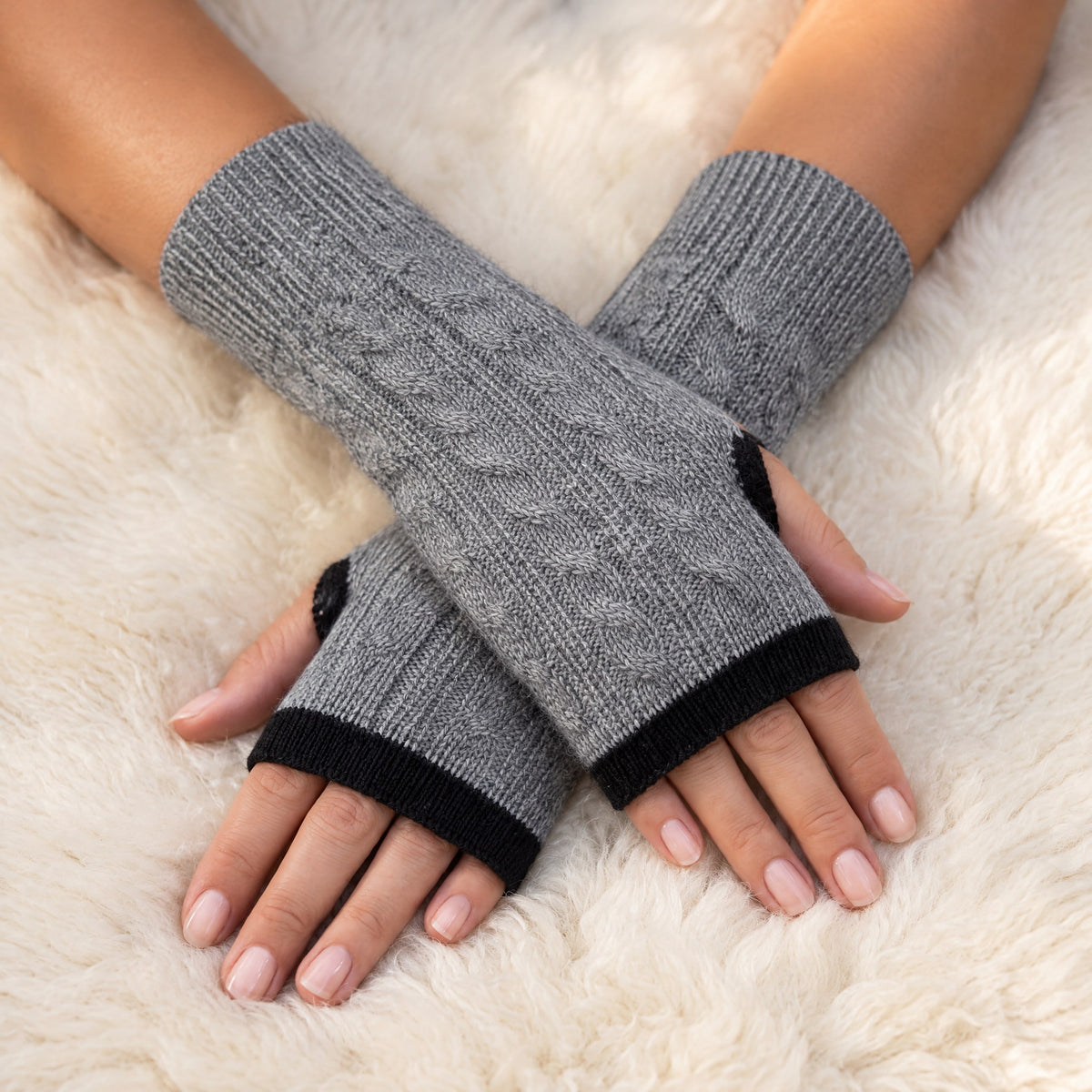 Grey & Black Reversible Cable Knit Baby Alpaca Fingerless Gloves worn on hands resting on a sheepskin rug