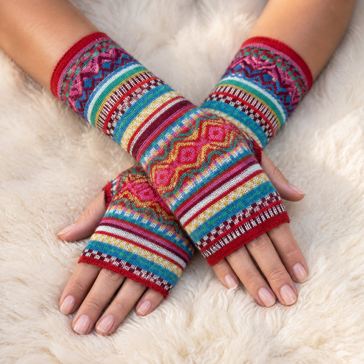 Red Rainbow Fair Isle Baby Alpaca Fingerless Gloves worn on hands resting on a sheepskin rug